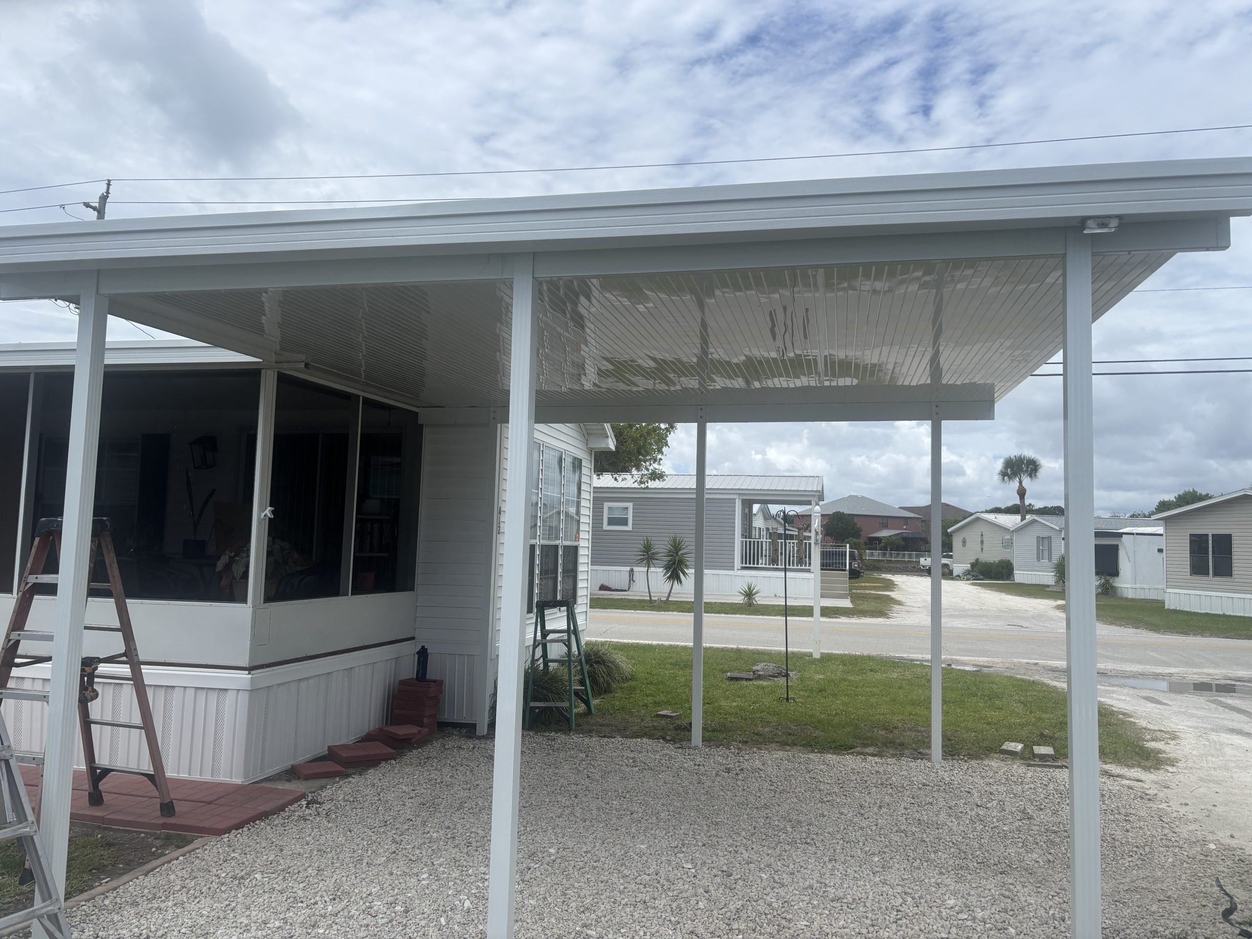 White aluminum 18-foot by 16-foot carport with ground-set posts beside an existing screen room at a Merritt Island, Florida mobile home park, sized for two compact vehicles